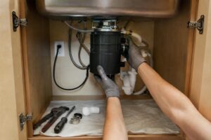Plumber installing a garbage disposal unit under a stainless steel kitchen sink in Oshawa